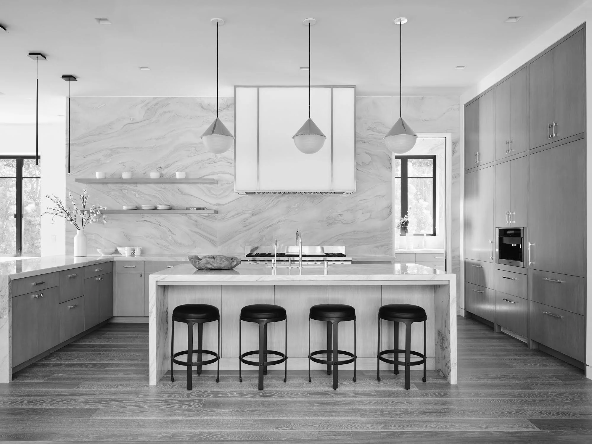Modern kitchen with light wood cabinets, marble countertops, and three hanging pendant lights over an island with four black stools. Open shelving with white dishes, a vase with flowers, and a window providing natural light.