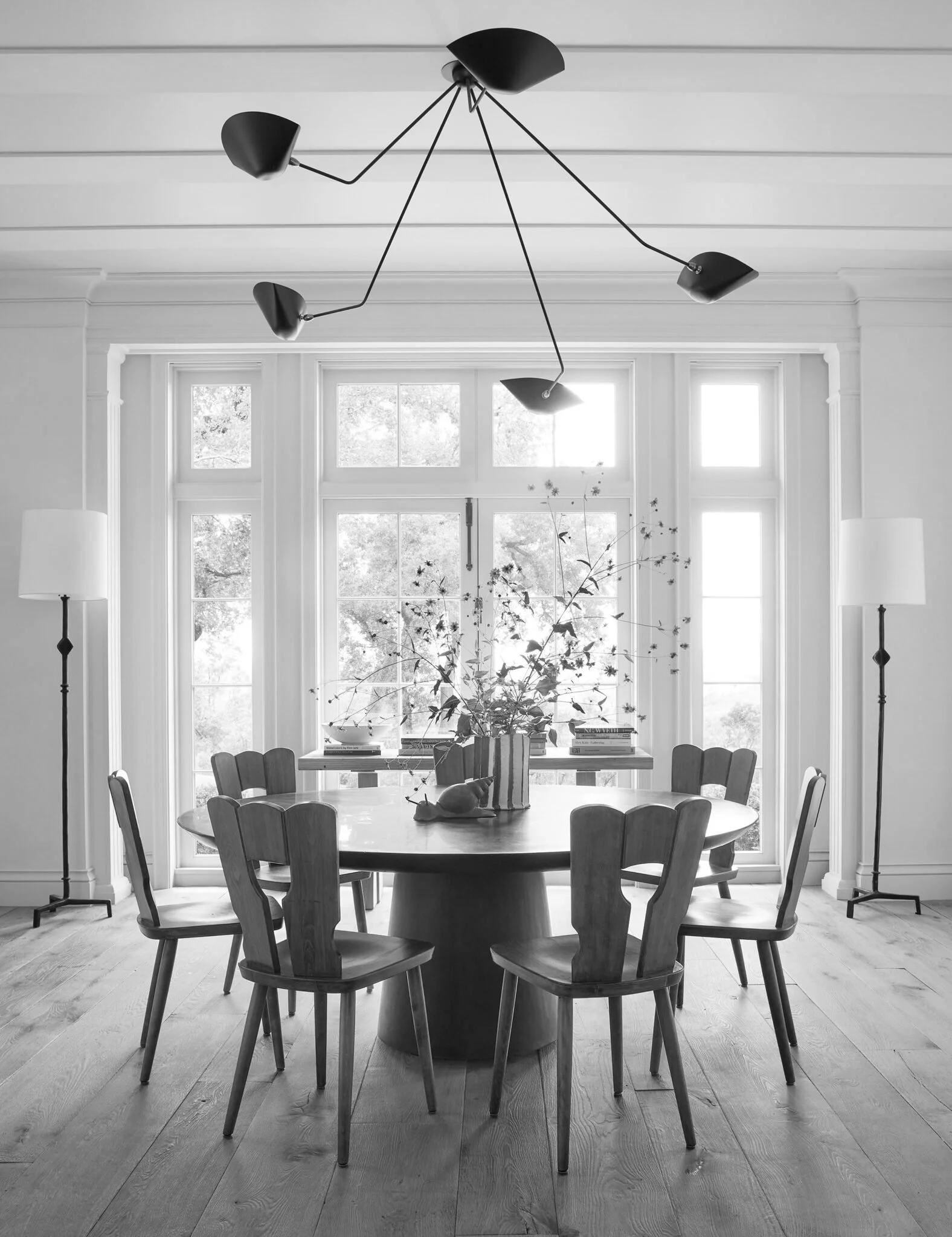 A black and white photo of a dining room with a round table, eight chairs, a large window with natural light, a table centerpiece with a vase and plant, books, and a modern chandelier with multiple black shades hanging from the ceiling.