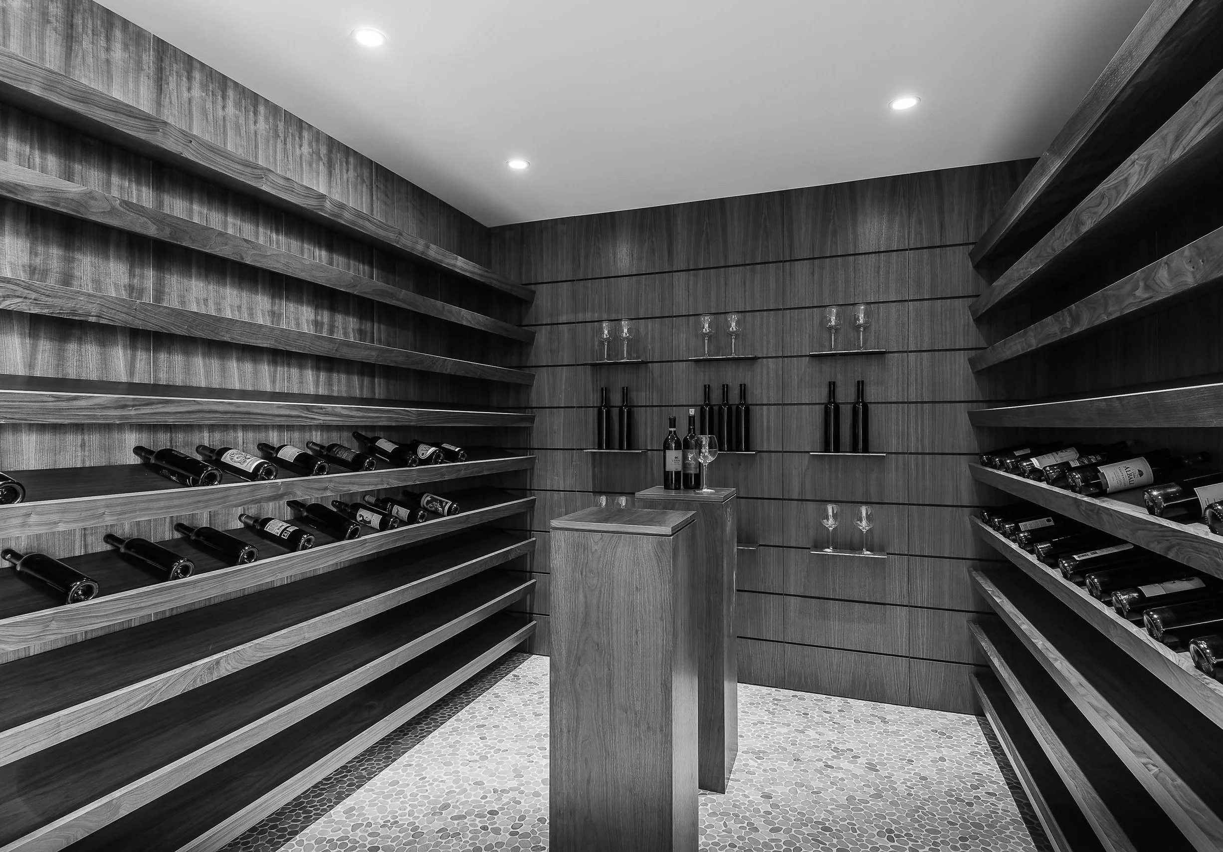 A wine cellar with wooden shelves containing bottles of wine, glasses, and wine bottles displayed on pedestal tables.