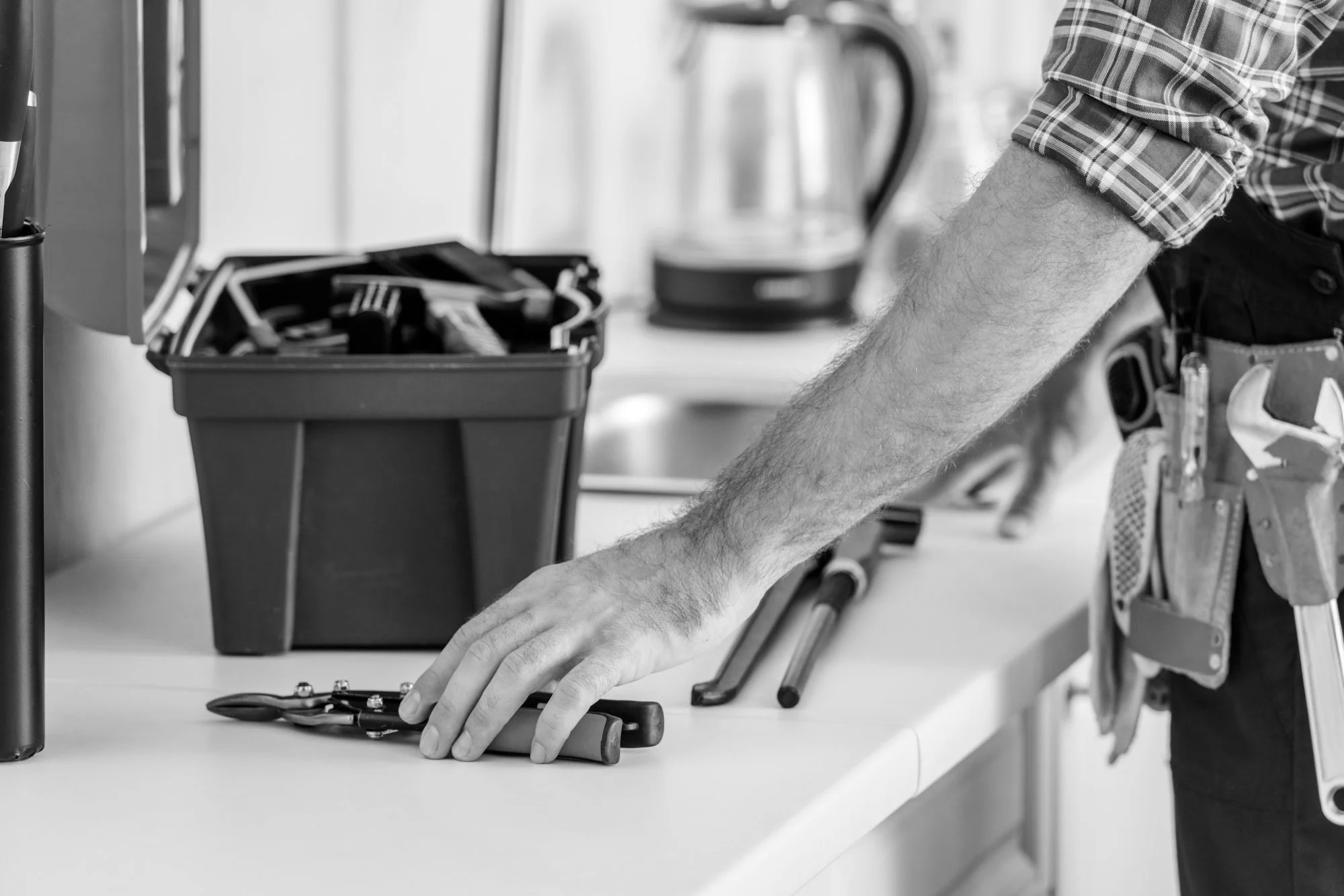 A person wearing a plaid shirt and a tool belt standing at a workbench, reaching for a wrench, with a toolbox and various tools visible on the workbench.
