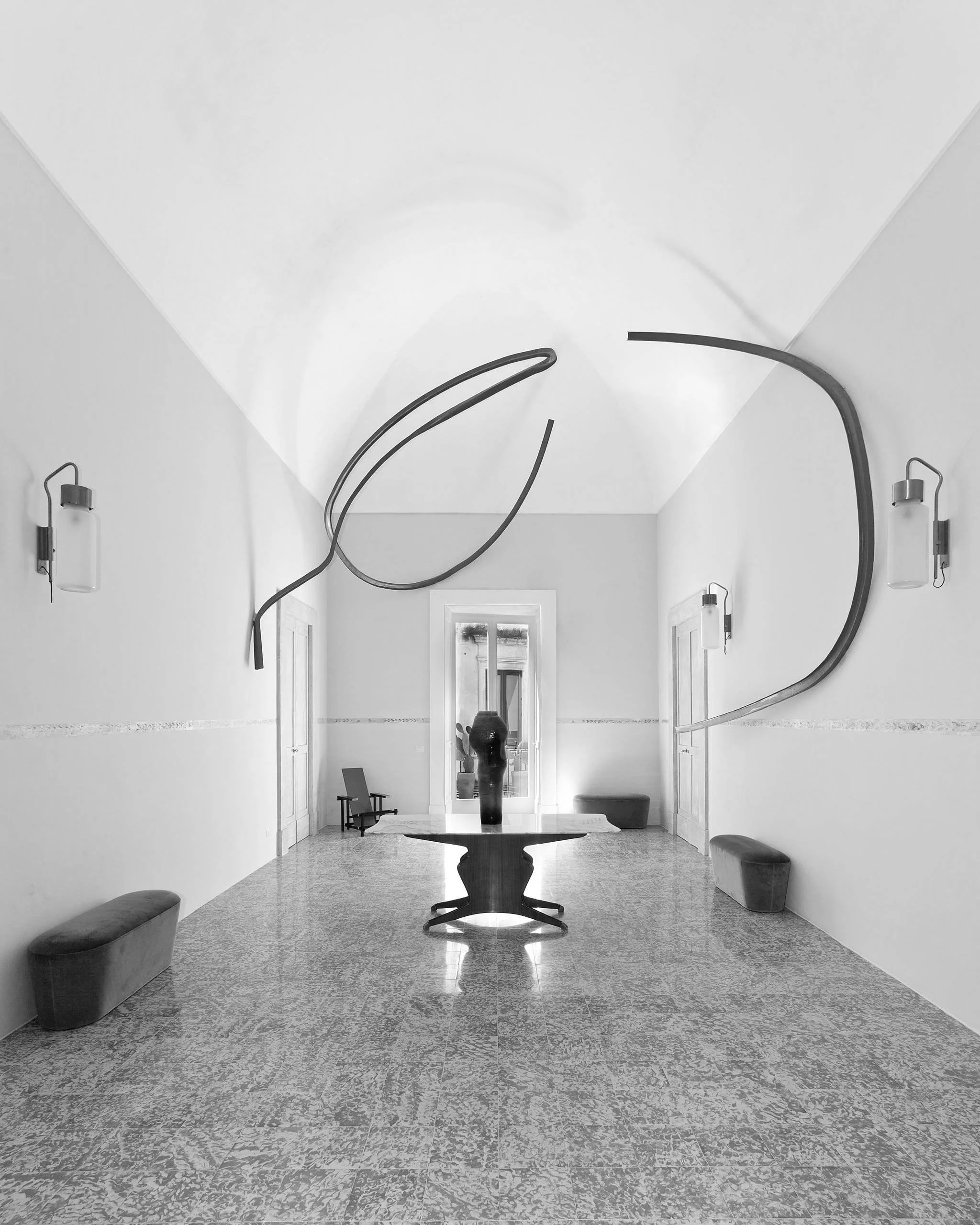 Black and white photo of a minimalist interior hallway with a wooden table, a sculpture, and curved metal wall art.
