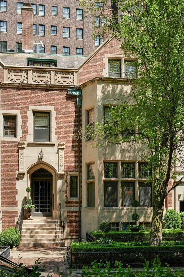 A historic brick and stone row house with front steps, a decorative entryway, and multiple windows, surrounded by greenery, in an urban setting.