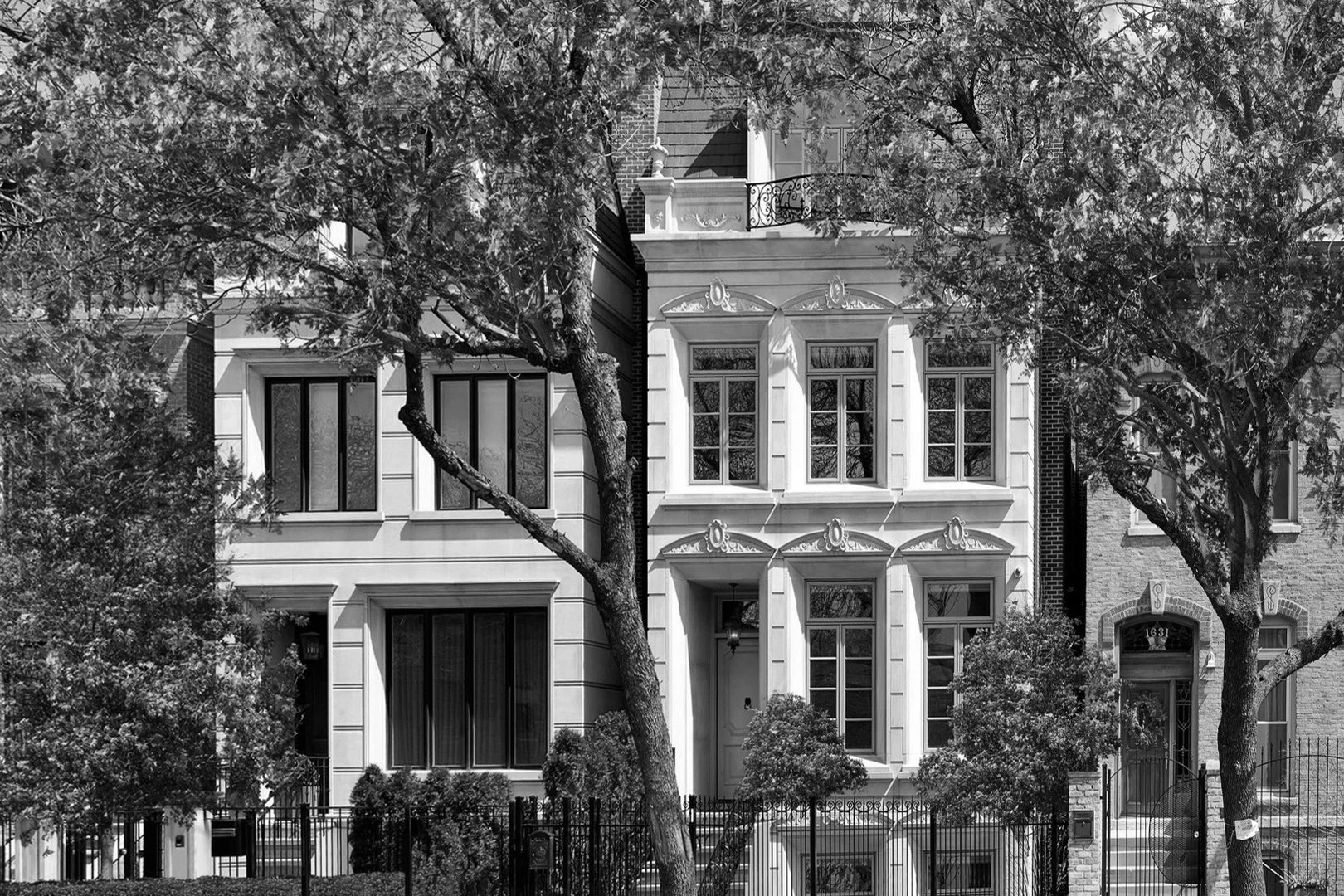 A black and white photo of a multi-story residential building with ornate architectural details, surrounded by trees and a wrought iron fence in the foreground.