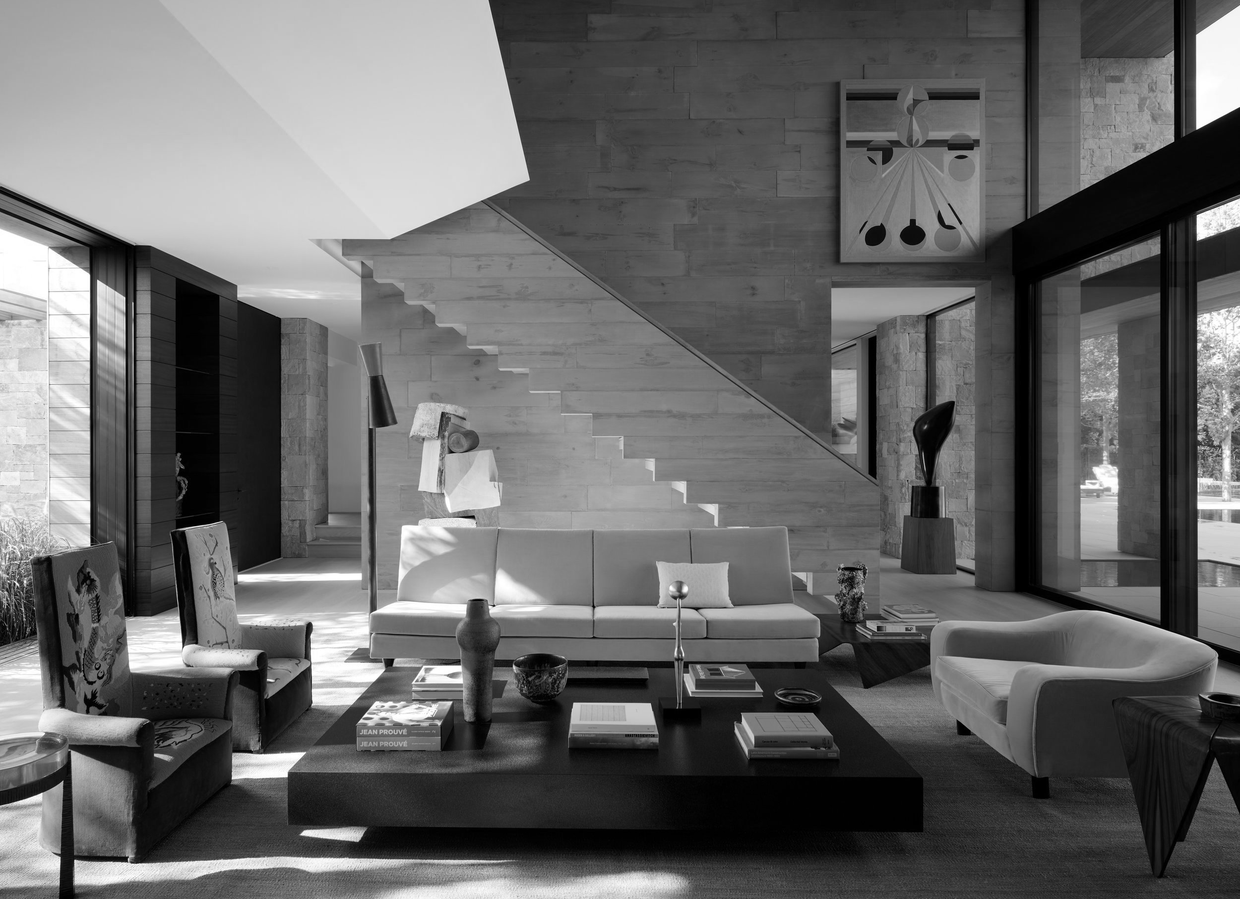Modern living room with a large white sofa, two patterned armchairs, and a black coffee table with books and decorative items. There are sculptures, large windows, and a staircase leading upstairs. Abstract art hangs on the wall.