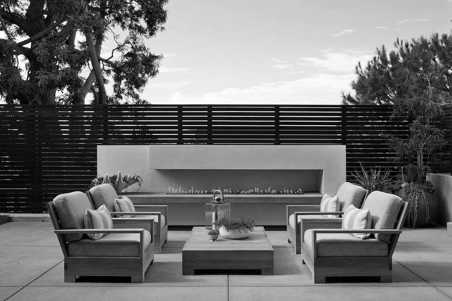 A modern outdoor patio with four cushioned armchairs arranged around a square wooden table, a fireplace built into a wall, and planters with plants, all enclosed by a horizontal slat wooden fence and surrounded by trees.