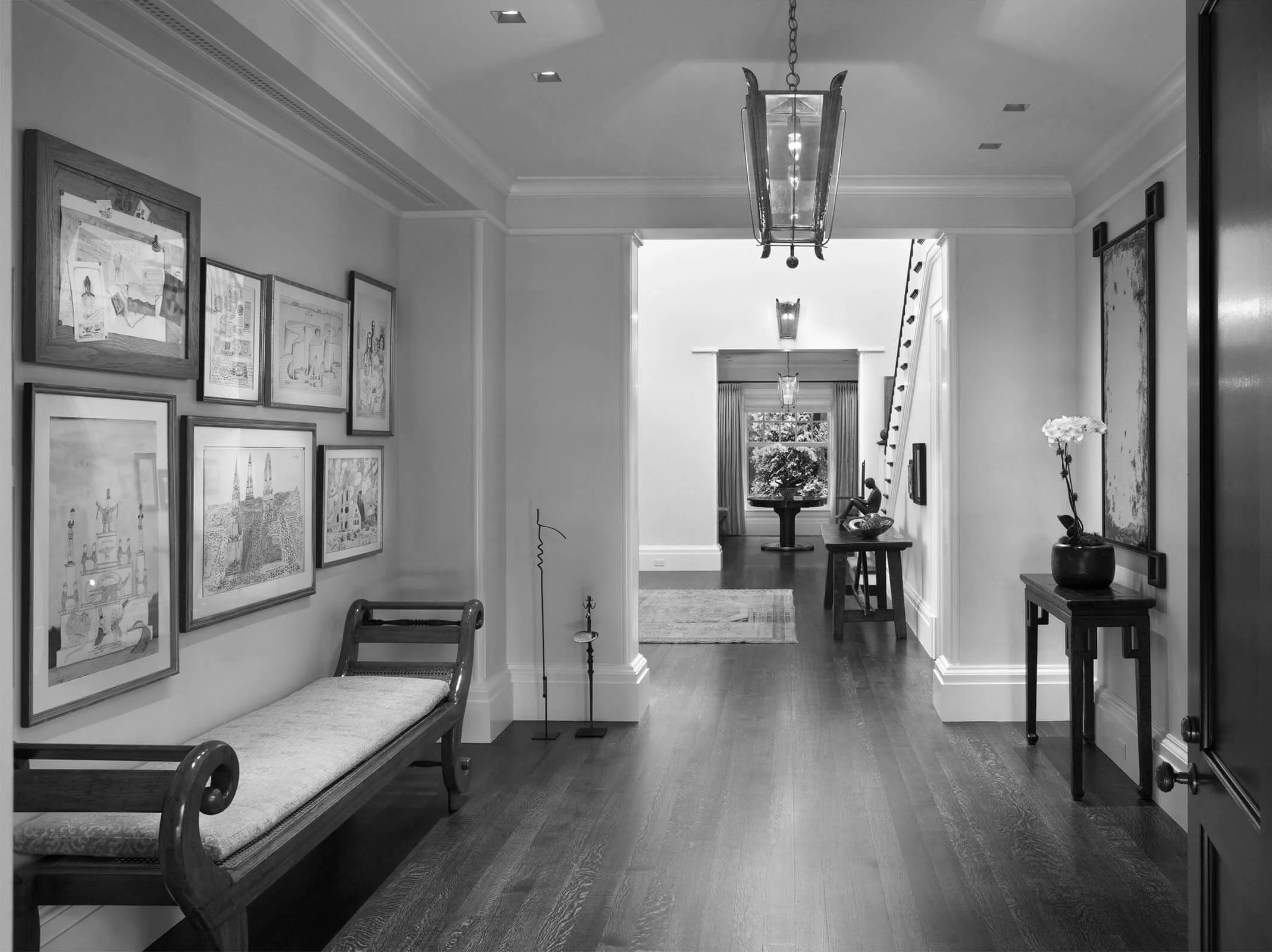 A black and white photo of a spacious hallway in a house, featuring framed artwork on the left wall, a wooden bench under the artwork, and a small table with a potted flower on the right. The hallway leads to a room with a window and a small table with a sculpture, with a staircase on the right side.