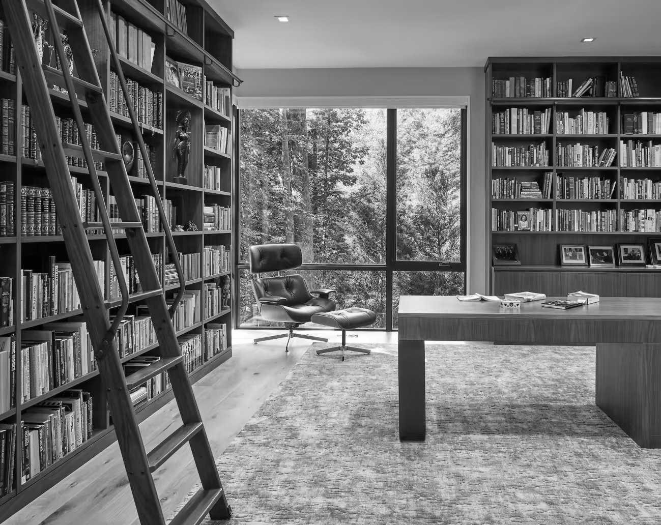 A room with large windows showing trees outside, filled with bookshelves, a lounge chair, a wooden desk with books, and framed photographs on the shelf.
