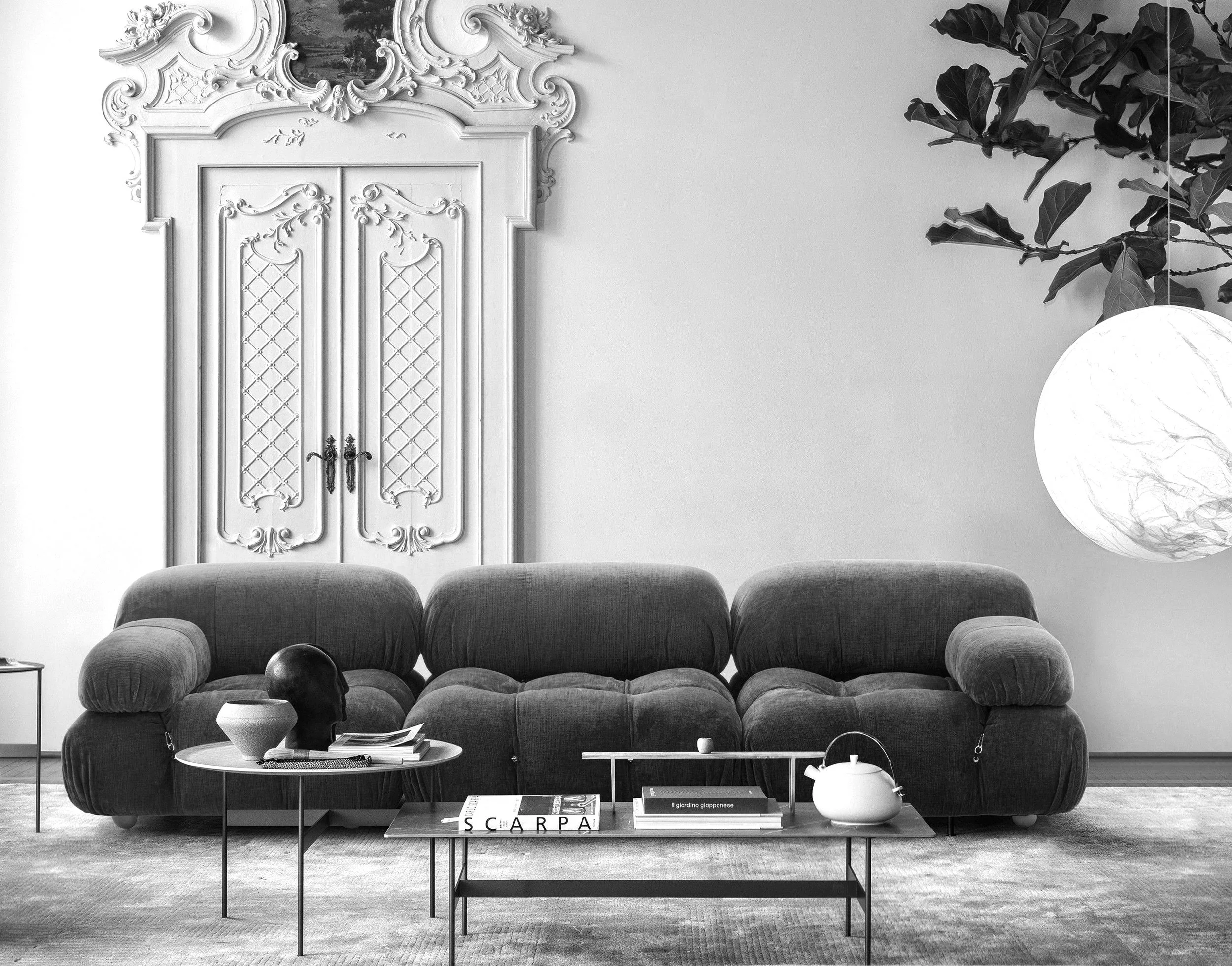 A modern living room with a dark textured sofa, a small round side table with decorative objects including a manikin head and books, and a rectangular coffee table with books, a teapot, and a small sphere. A large potted plant hangs over a glowing paper lantern on the right, with intricate carved white double doors with ornate trim and decorative details in the background.