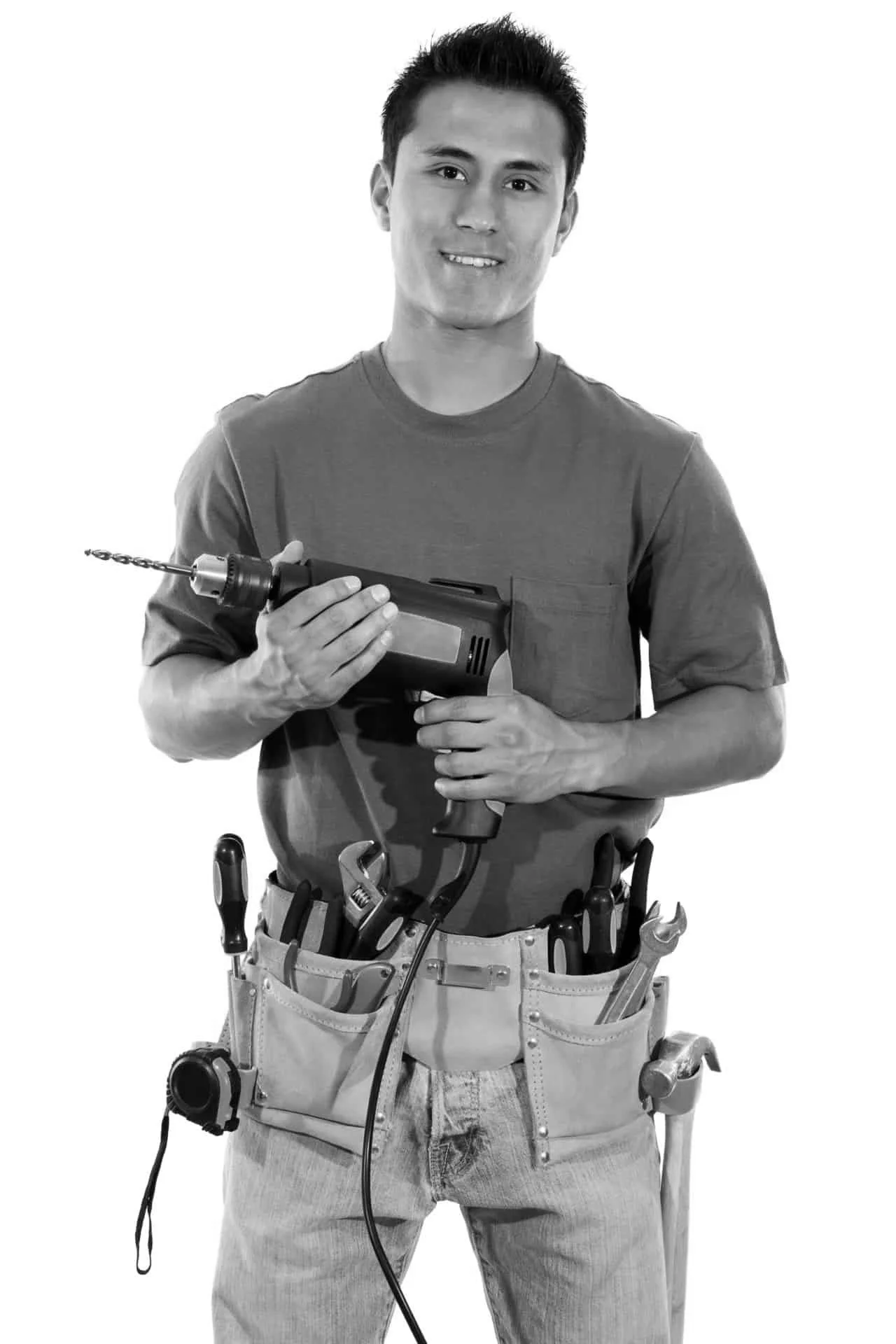 A young man wearing a utility belt filled with tools, holding a power drill with a drill bit, smiling at the camera.