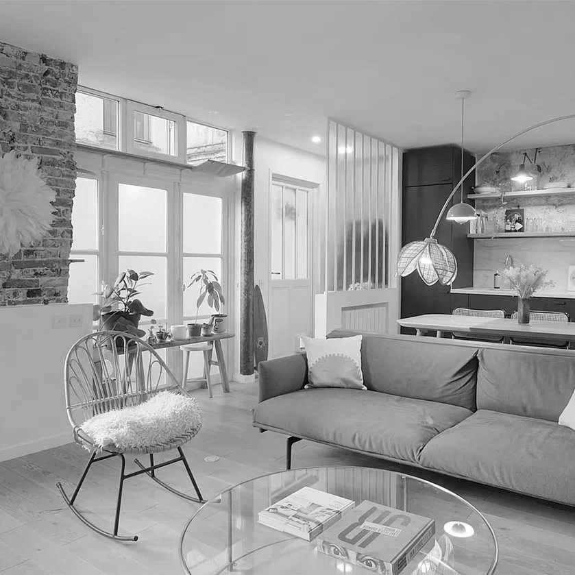 A modern living room with a sofa, a rocking chair with a sheepskin cushion, a glass coffee table, a large window with potted plants, and a dining area with hanging pendant lights.