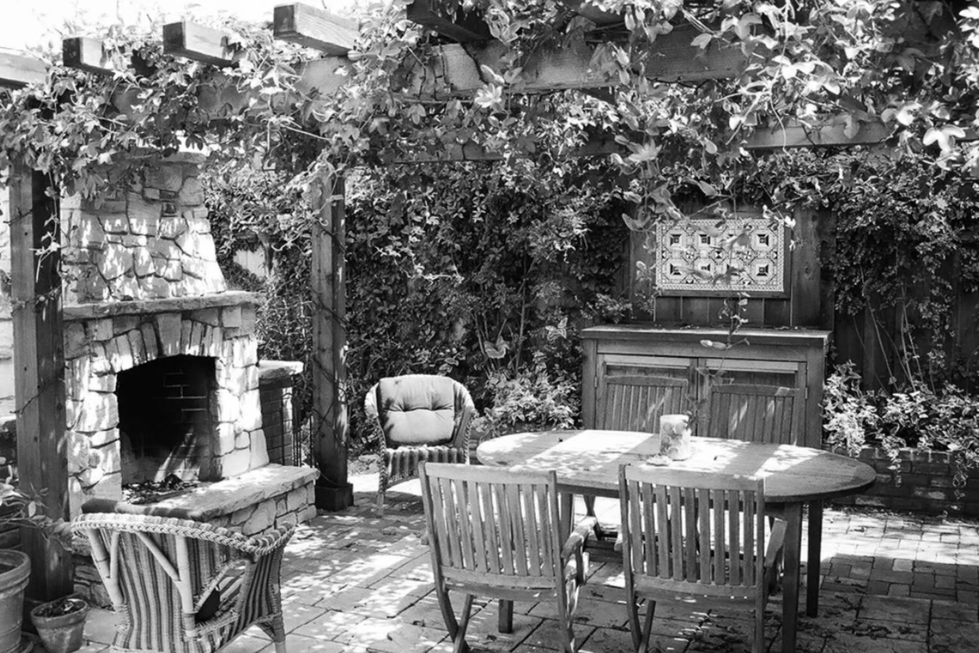 Outdoor patio with a stone fireplace, wooden dining table with chairs, cushioned armchair, wooden cabinet, decorative wall hanging, and lush foliage in the background.