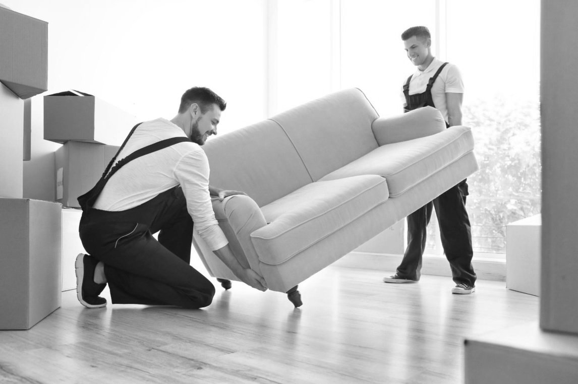 Two men moving a sofa in a room with sunlight, surrounded by cardboard boxes.