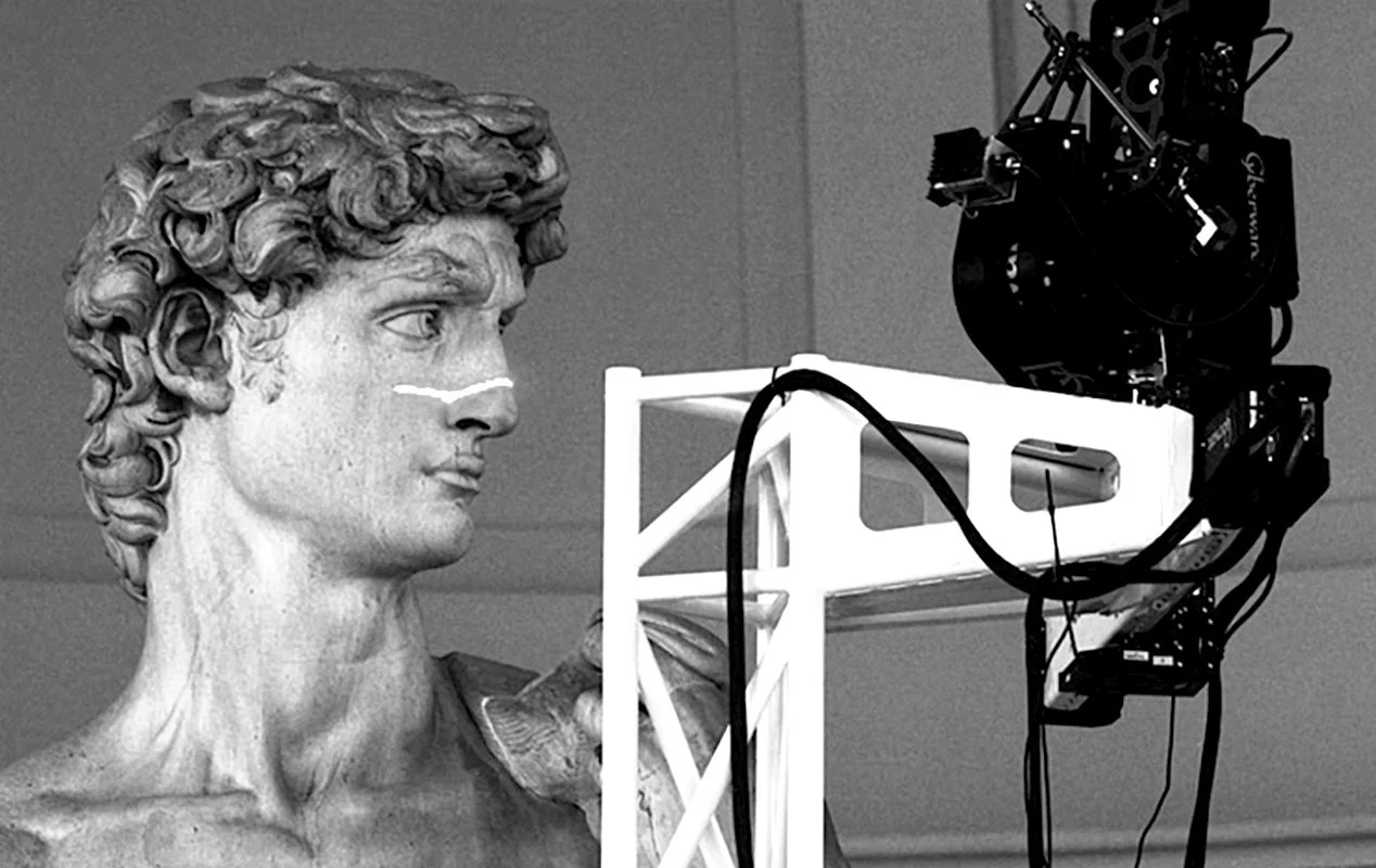 Photograph of a classical marble bust sculpture of a man's head and shoulders, facing left, in front of a black camera and white metal stand with wires.