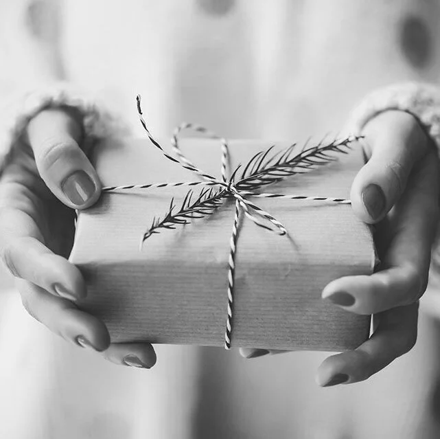 Person holding a wrapped gift tied with Christmas-themed twine and decorated with a sprig of pine.