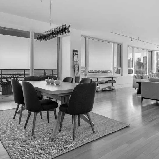 Modern dining and living area in an apartment with large windows, a dining table with six chairs, a chandelier, and a sofa, all in black and white.
