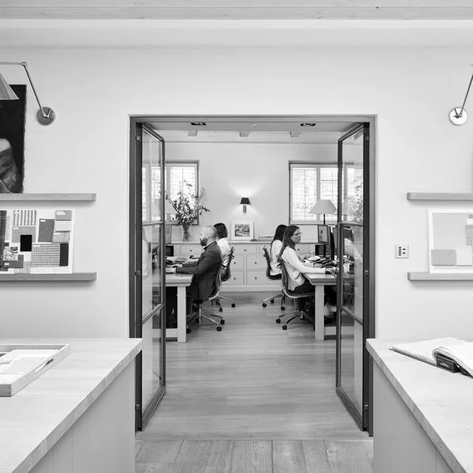 Office workspace with four individuals working at desks, two men and two women, viewed through open glass doors in a modern, well-lit office with plants and windows.