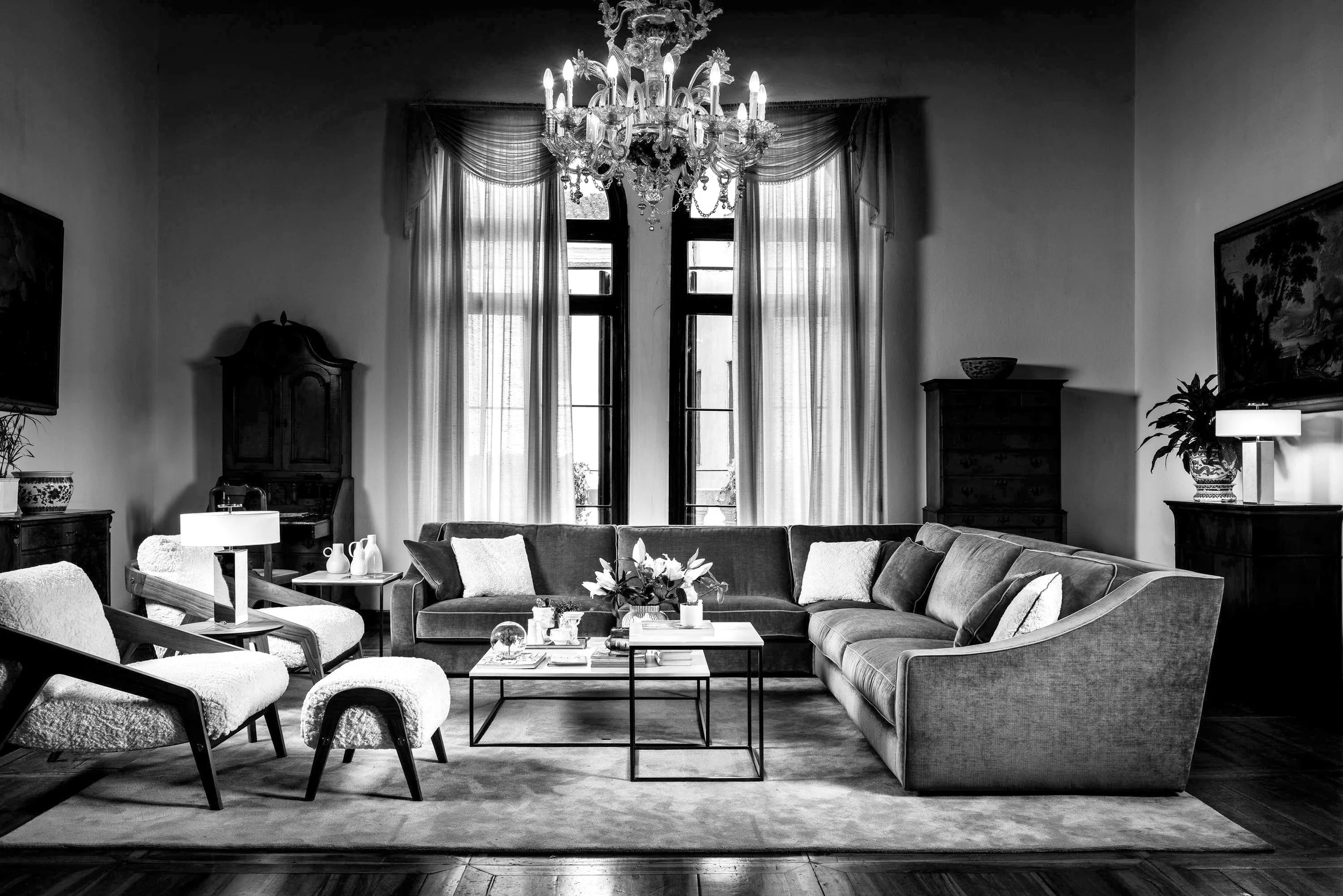 A black and white photo of a spacious living room with a large sectional sofa, armchairs, and a chandelier hanging from the ceiling. Tall windows with curtains let in light, and there are tables, lamps, and decorative items throughout the room.