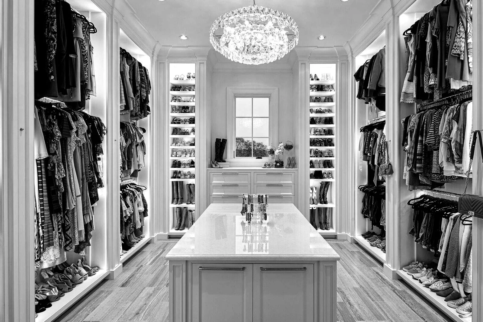 A luxurious walk-in closet with white shelving units filled with shoes, handbags, and accessories. There is a central island with a marble top, a window at the back with a view outside, and a decorative chandelier hanging from the ceiling.