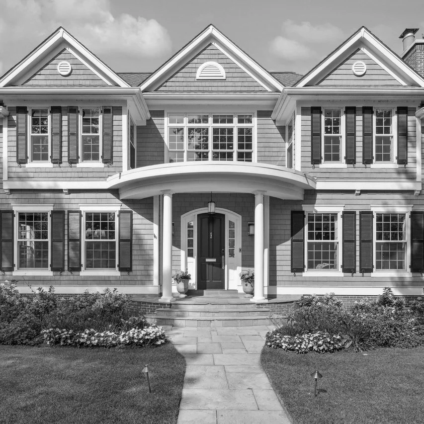 Black and white photo of a two-story house with a front porch, columns, multiple windows with shutters, and a landscaped front yard with flowers and grass.