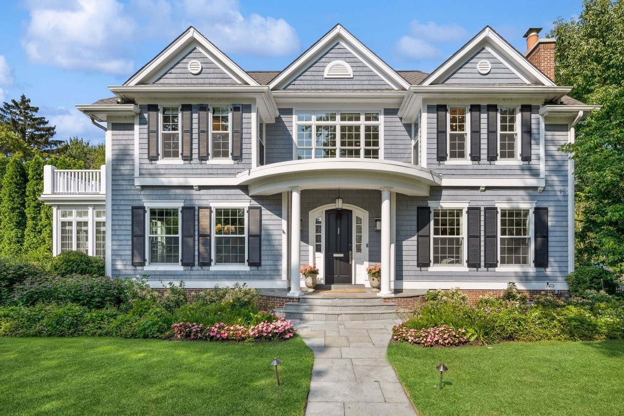Front view of a large, two-story house with gray siding, black shutters, and a black front door, surrounded by a landscaped yard with pink flowers and green grass.