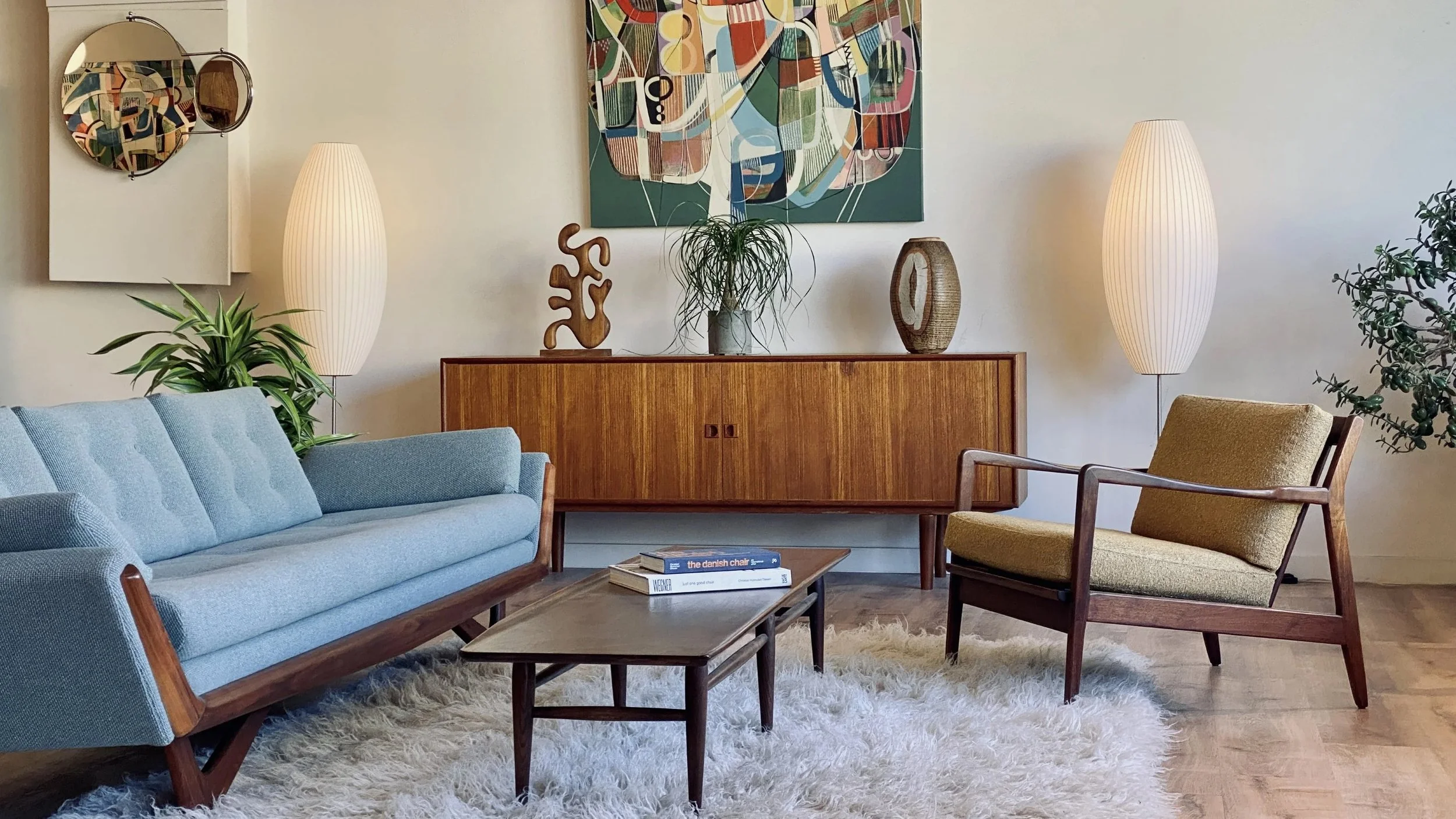 Living room with a light blue sofa, a wooden coffee table with books, a beige armchair, and a wooden sideboard with decorative items and greenery. There are two tall, white floor lamps and abstract artwork on the wall.