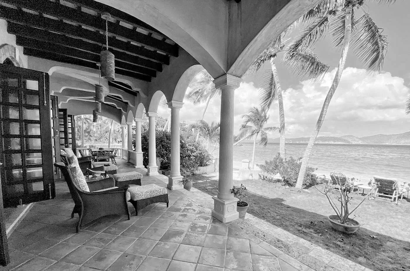 A porch with tiled flooring and arched columns overlooking a beach with palm trees and a body of water, with lounge chairs and potted plants.