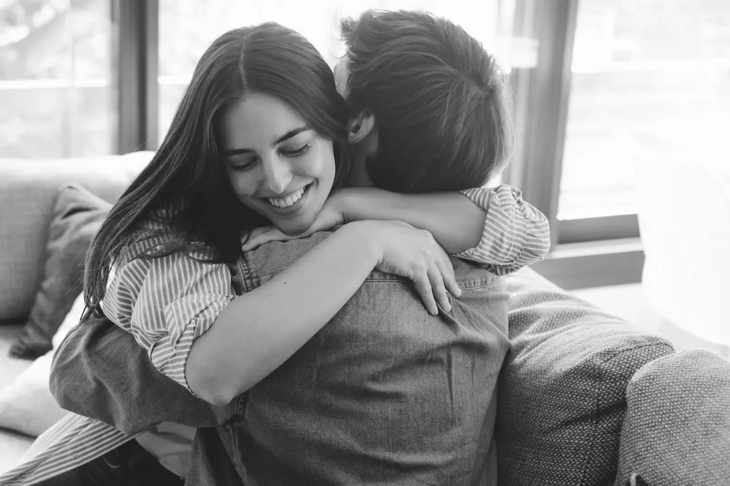 A woman smiling with her eyes closed hugging a person who is facing away, in an indoor setting with a window in the background, in black and white.