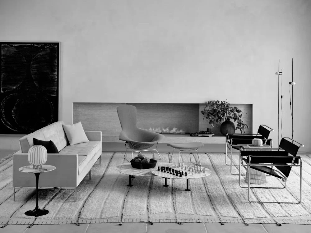 Modern living room with white sofa, black pillows, contemporary chair, coffee table with chess and decorative bowl, striped rug, fireplace with plant, two black chairs with side table, and hanging pendant lights.