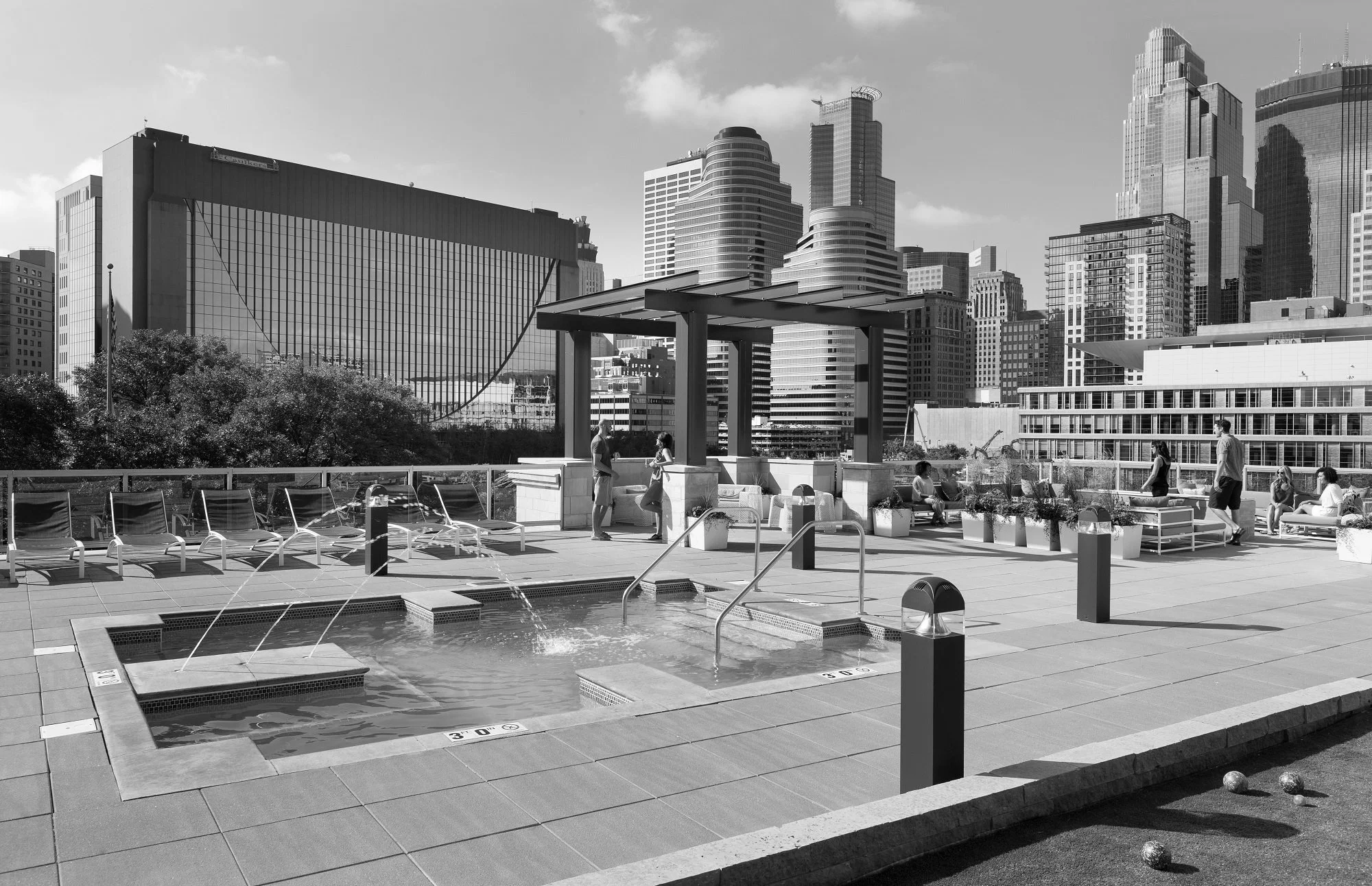 Rooftop pool with lounge chairs, hot tubs, and people relaxing, surrounded by city skyscrapers.