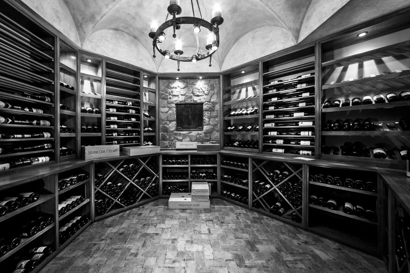 Black and white photo of a wine cellar with wooden shelves filled with wine bottles, a stone accent wall, and a chandelier hanging from the ceiling.