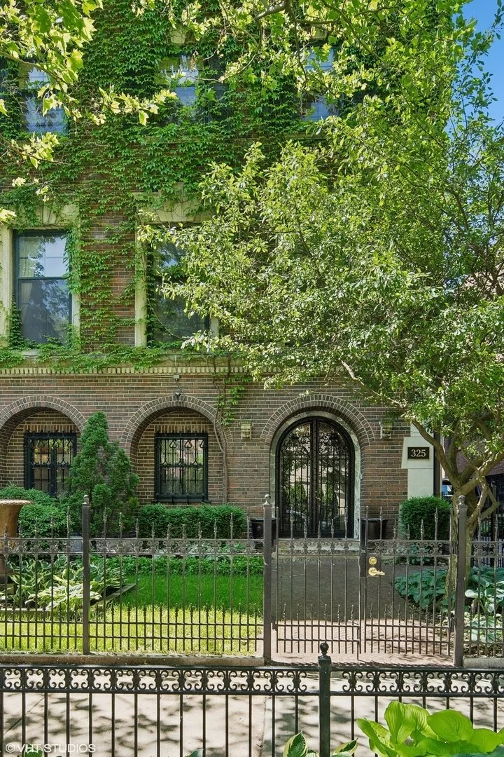 A brick residential building with arched windows and a black iron gate, surrounded by lush green trees and plants, with a sidewalk and a decorative fence in front.