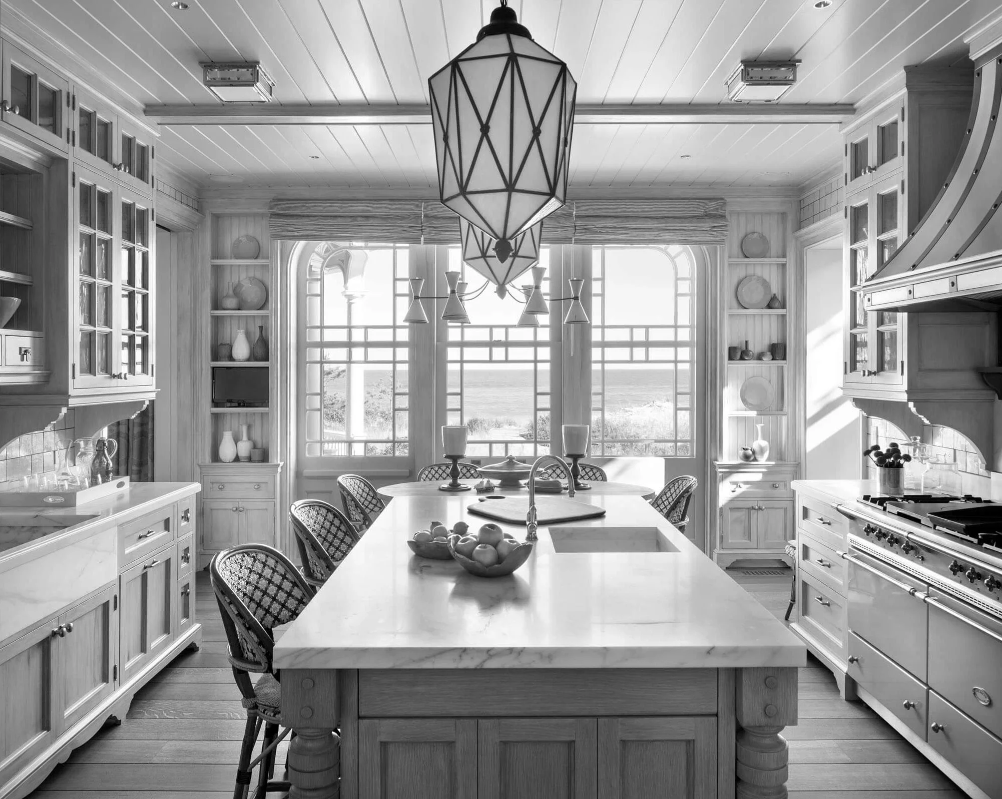 A black and white photo of a spacious kitchen with a large island in the center, surrounded by six chairs. The kitchen features wooden cabinets, shelves with decorative items, a stove, and a large window with a scenic view outside. A geometric pendant light hangs above the island.