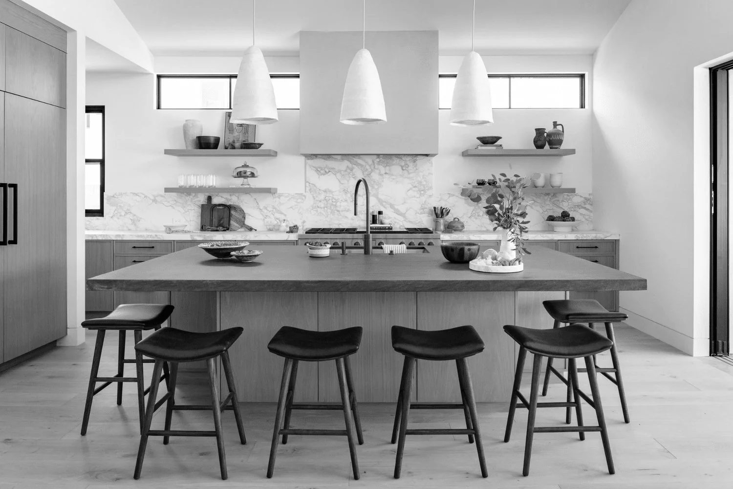 Modern kitchen with a large island, four bar stools, pendant lighting, marble backsplash, open shelves with dishes and decor, and a faucet over the sink in monochrome.