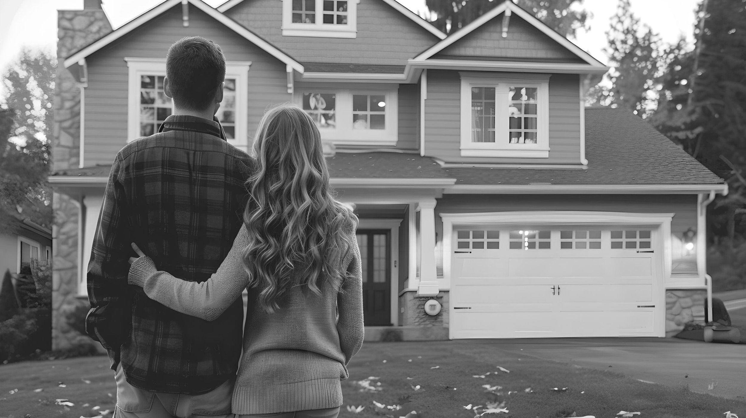 A man and woman stand close together in front of a large house, looking at it. The house has multiple stories, a garage with windows, and a front porch. The image is in black and white.