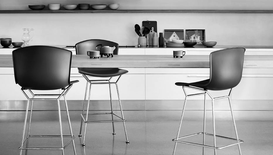 Black and white image of a modern kitchen with a wooden counter and three high chairs. There are bowls, cups, and decorative items on the counter, along with artwork and utensils in the background.