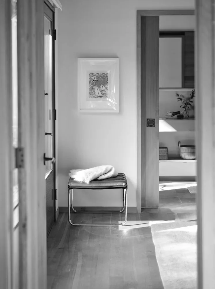 A black and white photo of a modern entryway with a bench, a fur throw, a framed picture on the wall, and a doorway leading to another room with a plant and shelves.