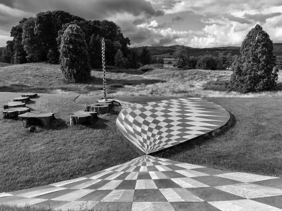 Black and white landscape with trees, hills, cloudy sky, and mural featuring checkerboard illusion on the ground and side of a hill.