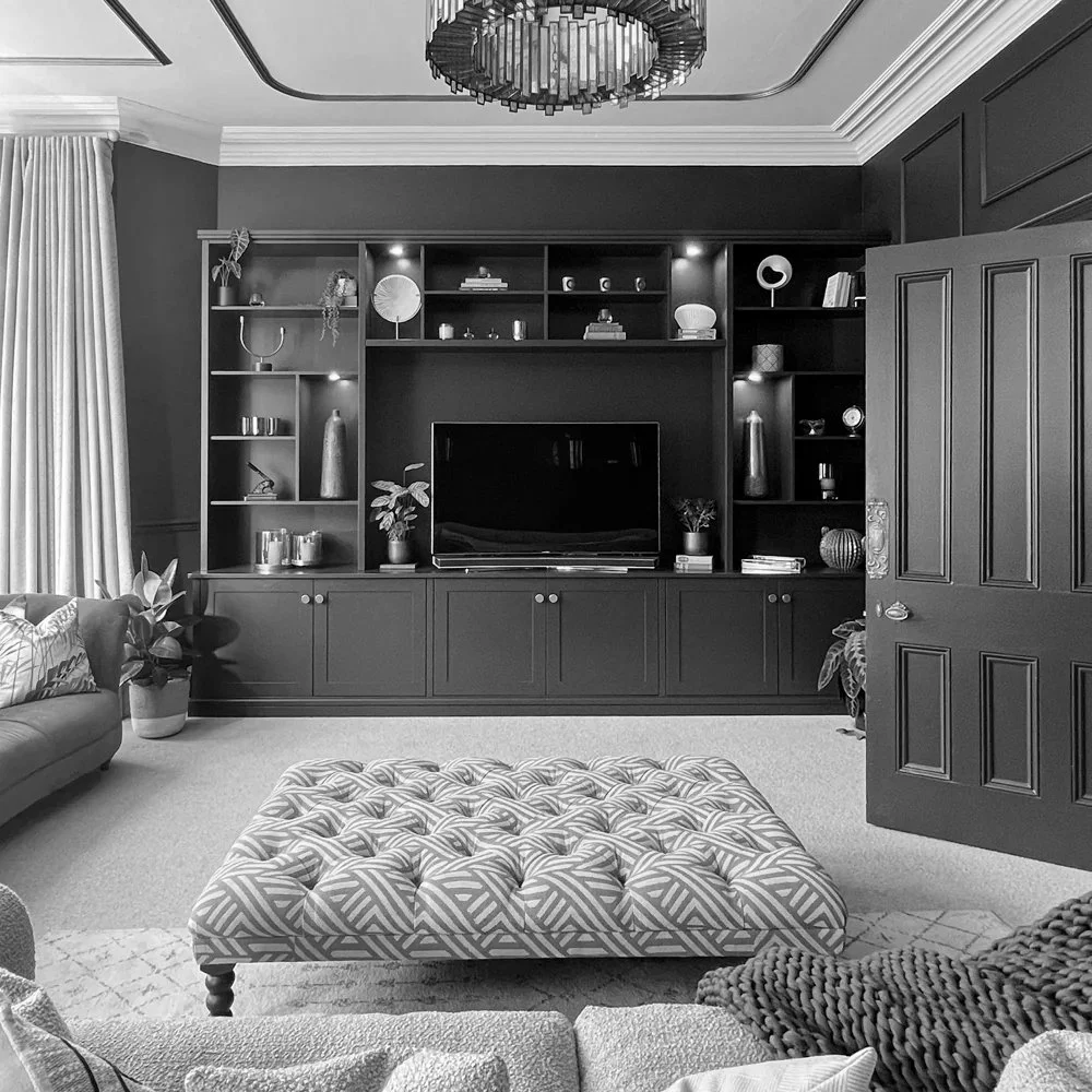A monochrome living room with a large built-in shelf unit holding decorative items and a flat-screen TV, a tufted ottoman in the center, a sofa with cushions, potted plants, a door ajar to the right, and a circular chandelier hanging from the ceiling.