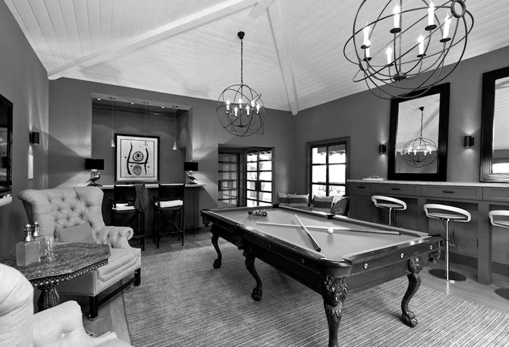 A stylish game room with a pool table, bar area with barstools, armchairs, modern chandeliers, and artwork on the walls in black and white.