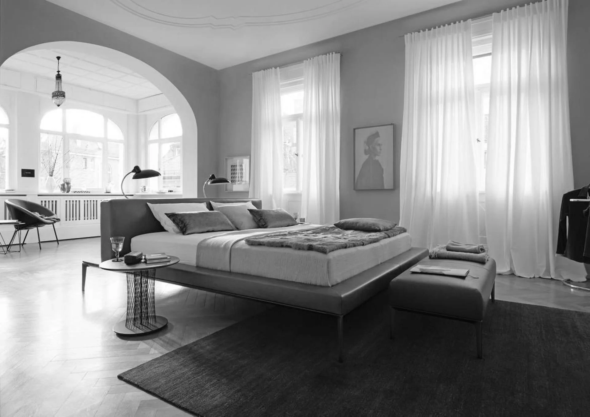 A modern, minimalist bedroom with a low bed, pillows, and a fur throw, surrounded by large windows with sheer curtains, and a small side table with a glass of water and stacked books.