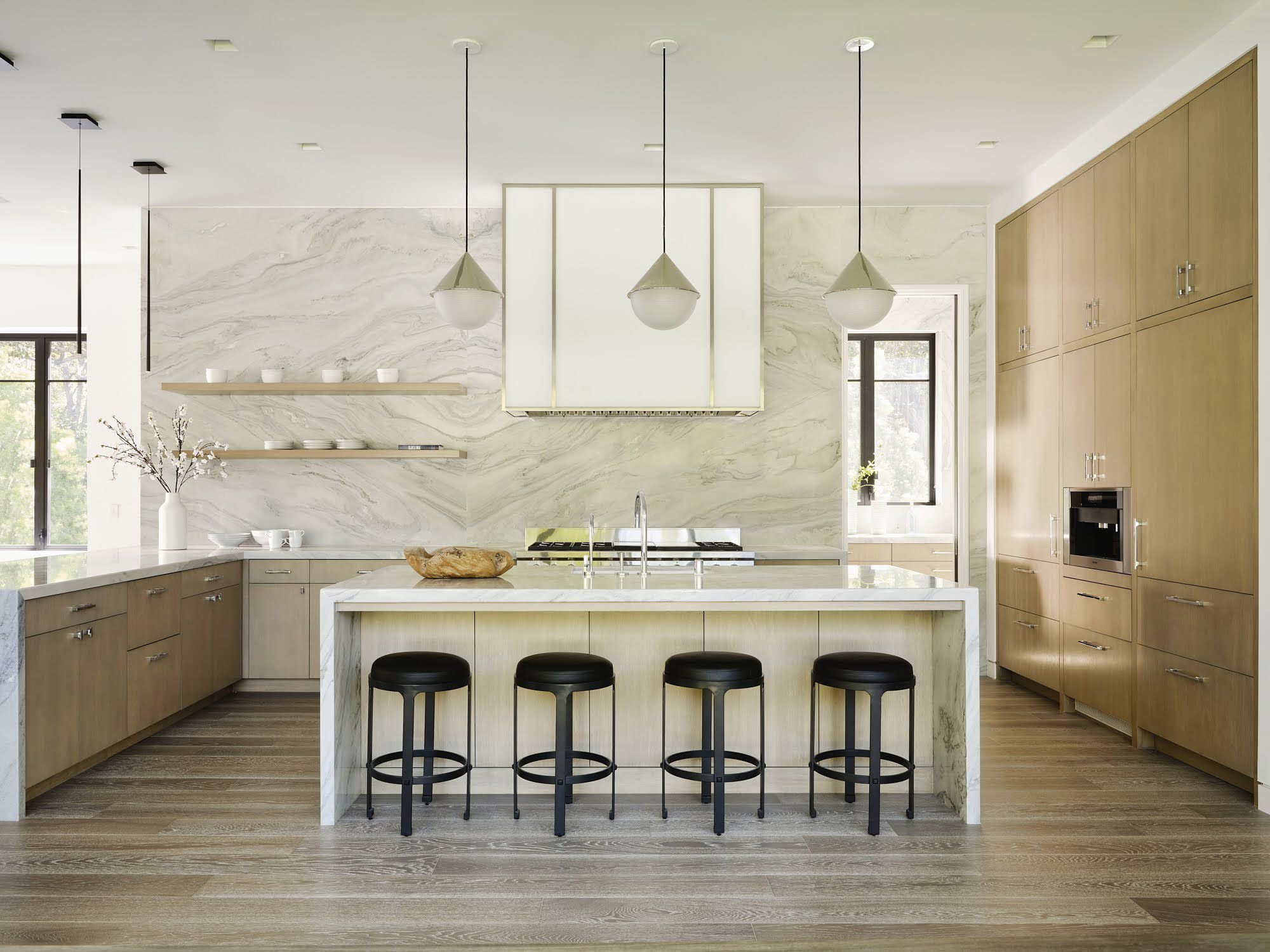 Modern kitchen with a marble backsplash, wooden cabinetry, a central island with a marble countertop, black bar stools, pendant lighting, and large windows.