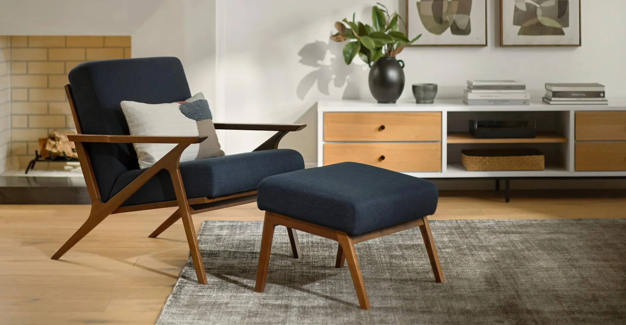 Modern living room featuring a black upholstered armchair with wooden armrests and matching ottoman, a sideboard with wooden and white drawers, a large black vase with greenery, framed abstract art on the wall, and a textured rug on light wood flooring.