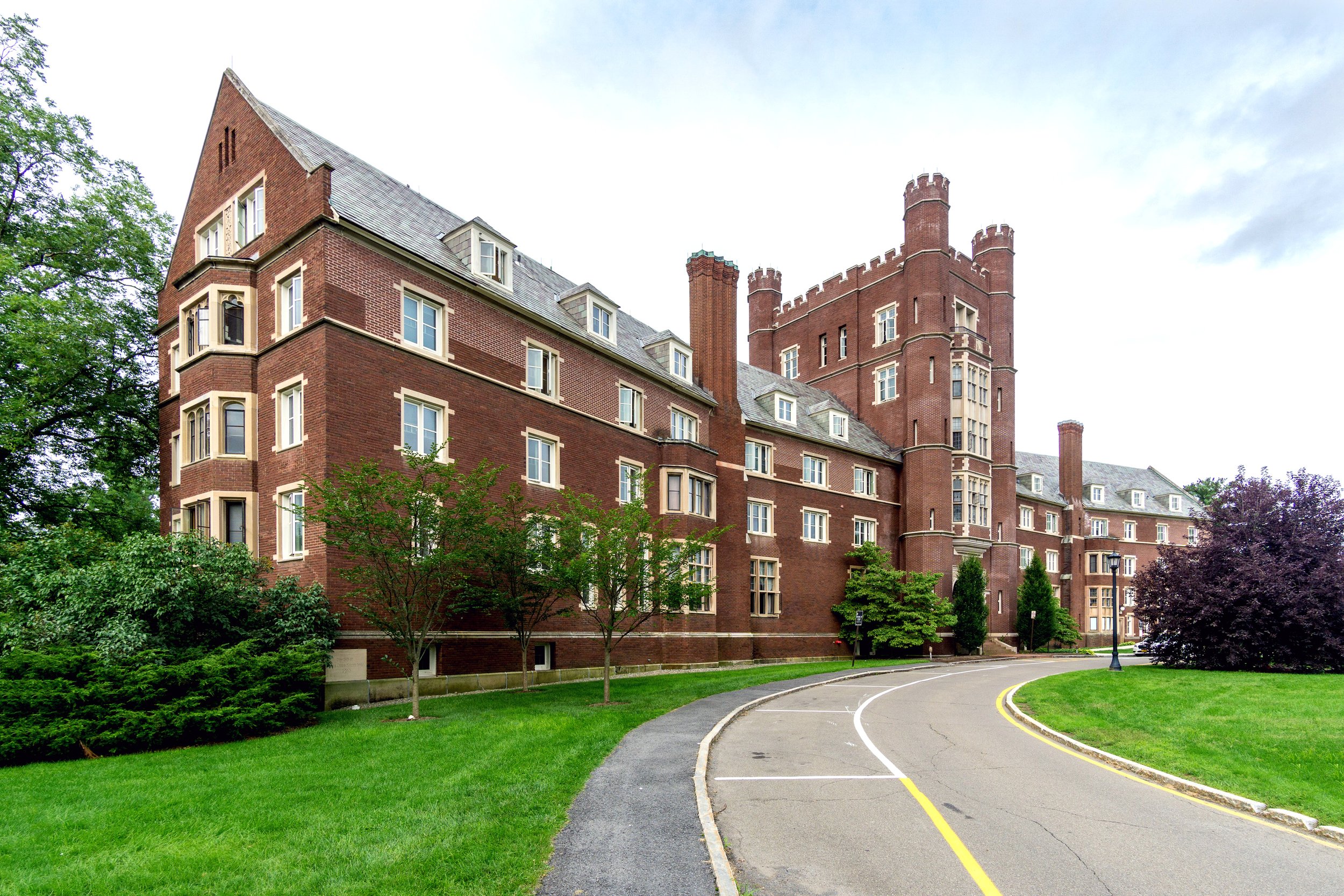 Red brick castle-style building with multiple floors, turrets, and chimney, surrounded by green lawn, trees, and a winding road in foreground under cloudy sky.