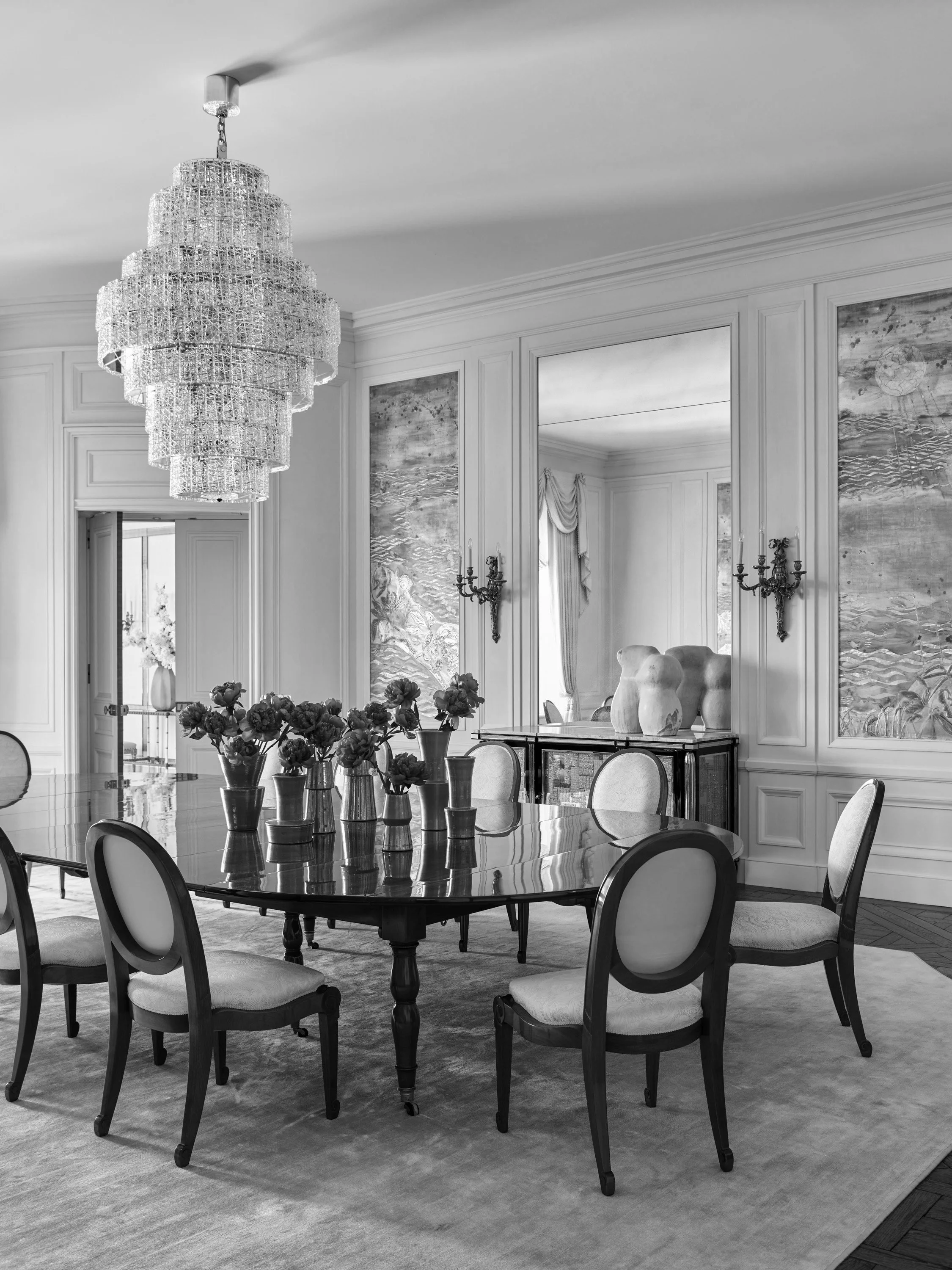 Elegant dining room with a large chandelier, a polished wooden table, and eight upholstered chairs. Decor includes vases with flowers on the table, large ceramic vases on a sideboard, wall sconces, and framed artwork on the walls.