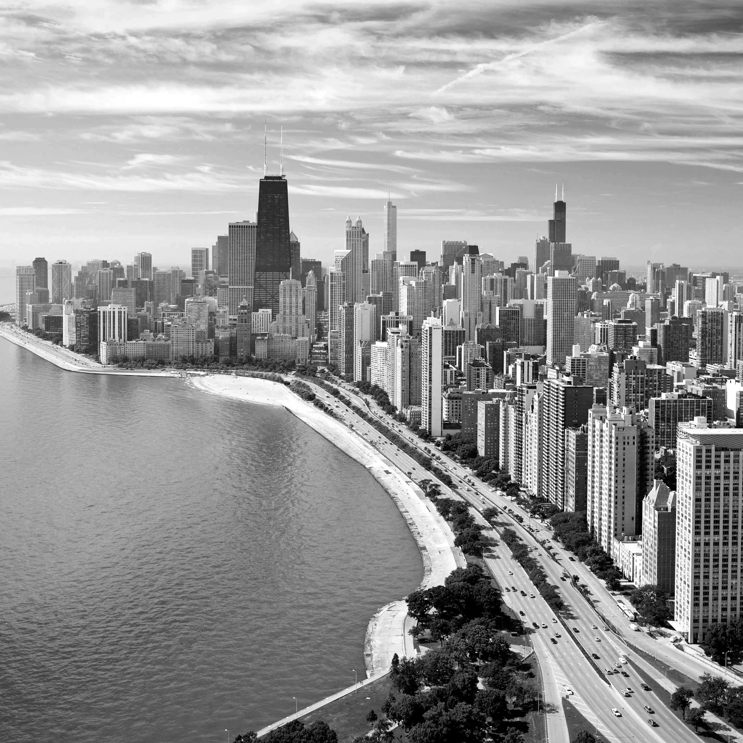 Black and white aerial view of Chicago skyline with Lake Michigan in foreground and skyscrapers, including Willis Tower, under a partly cloudy sky.