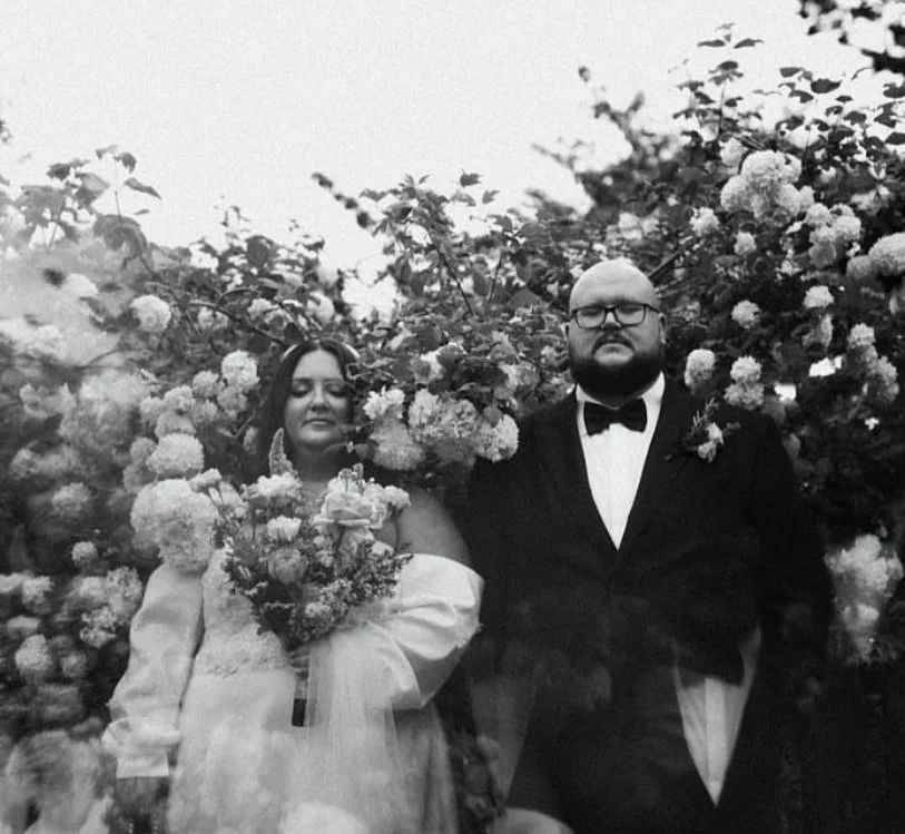 Black and white photo of a bride and groom standing in front of a large bush with many flowers. The bride is holding a bouquet and wearing an off-the-shoulder wedding dress, while the groom is in a tuxedo with a bow tie.