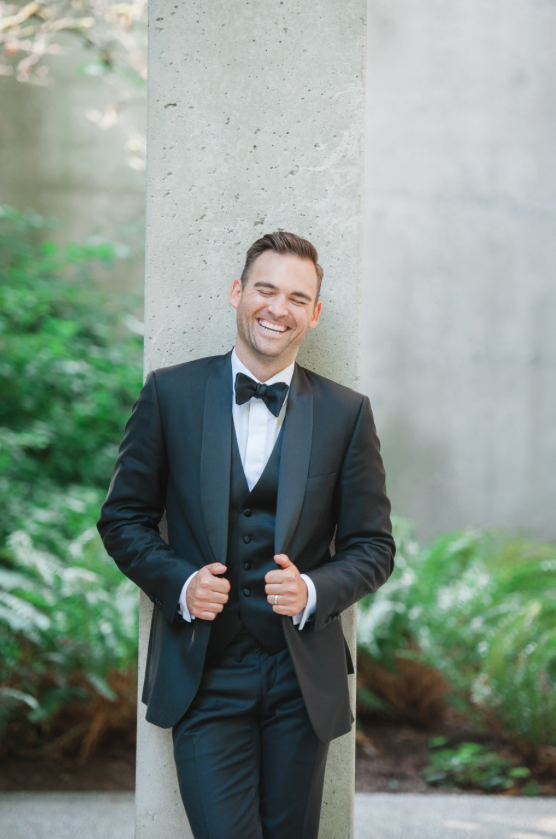 Man in tuxedo smiling and laughing outdoors, leaning against a concrete pillar