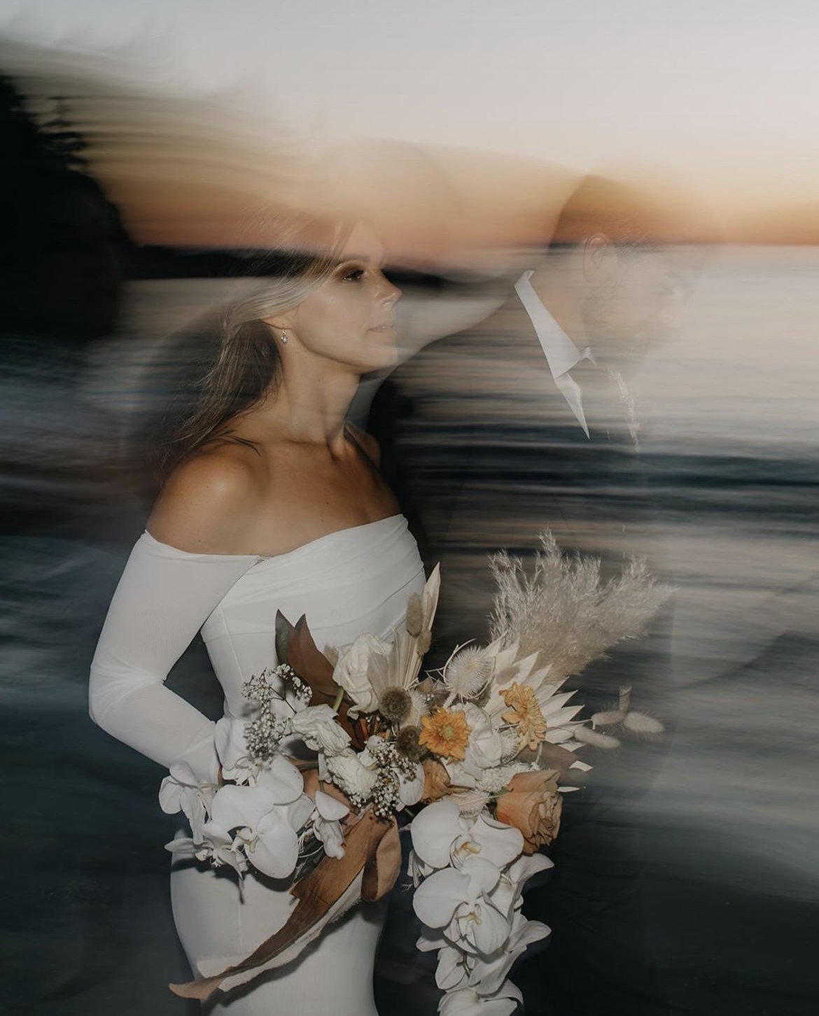 A blurred photo of a woman in a white off-shoulder dress holding a bouquet of white and beige flowers, with a sunset background and a man in a suit walking beside her, creating a motion effect.