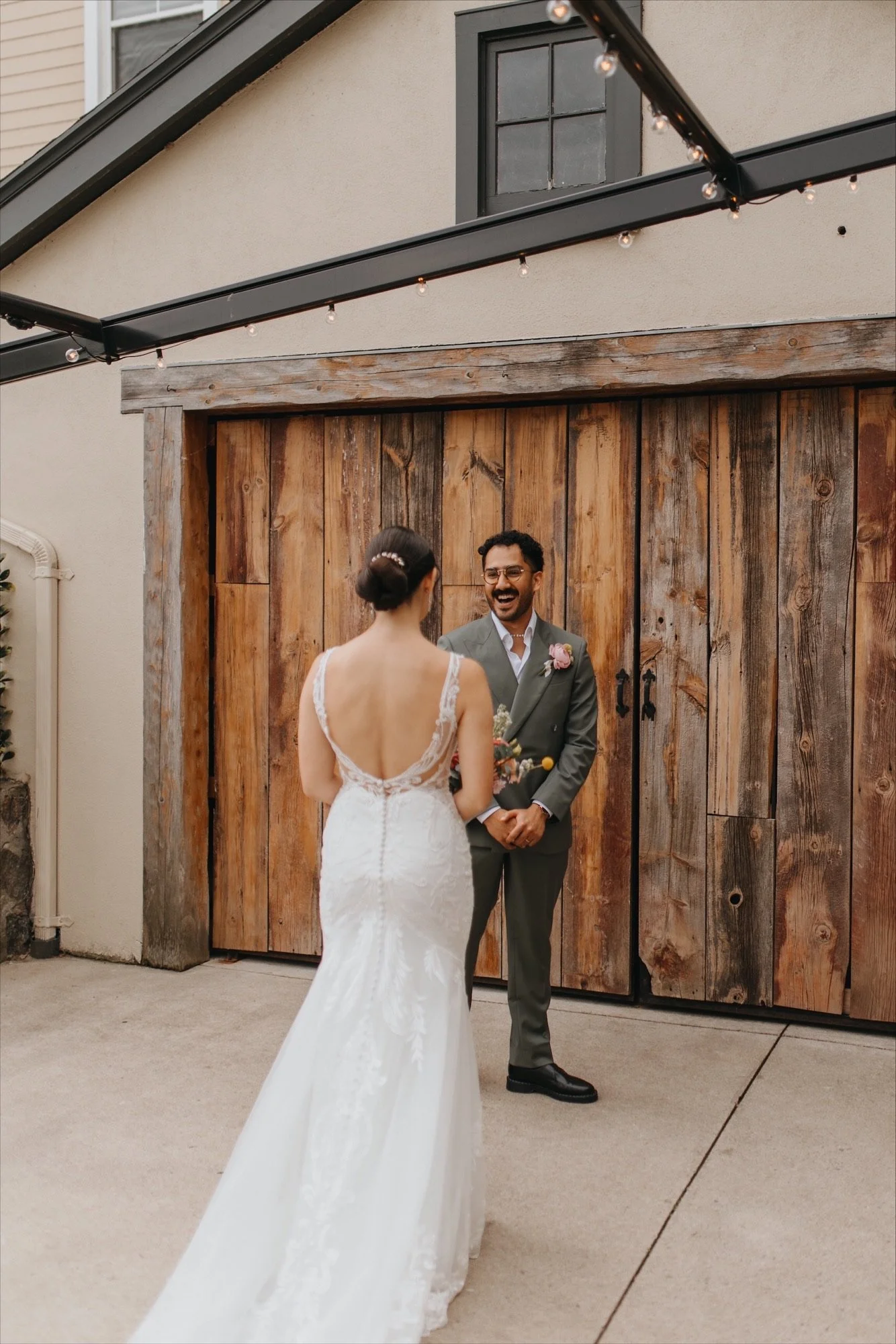 A bride and groom exchanging vows outdoors in front of a wooden barn door. The bride is wearing a white gown with a low back, and the groom is in a gray suit with a boutonniere, smiling at her.