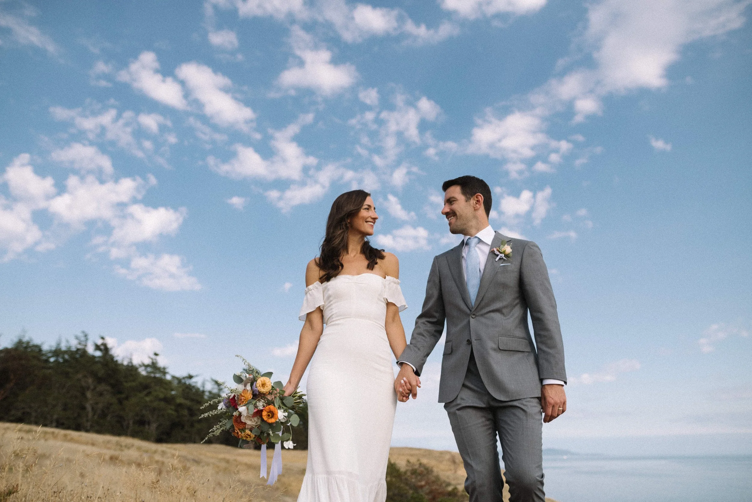 A bride and groom holding hands and smiling at each other outdoors on a cloudy day.